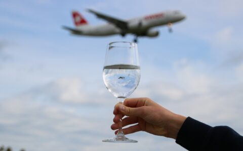 A hand holds a wine glass reflecting the sky, with a blurred airplane flying overhead