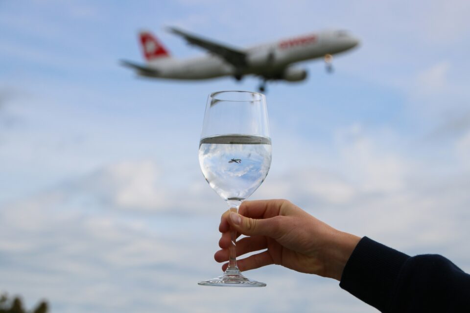 A hand holds a wine glass reflecting the sky, with a blurred airplane flying overhead