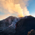 Active volcano erupting with glowing lava flows streaming down its dark slopes and a large ash cloud billowing above