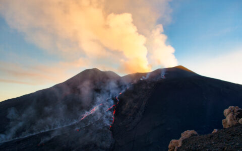 Active volcano erupting with glowing lava flows streaming down its dark slopes and a large ash cloud billowing above