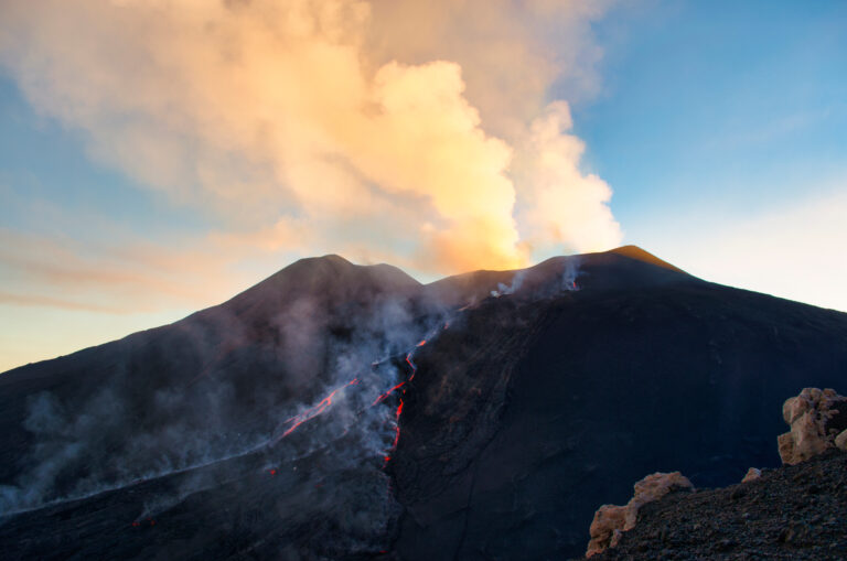 Active volcano erupting with glowing lava flows streaming down its dark slopes and a large ash cloud billowing above