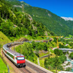 A red and white passenger train winds through a lush Alpine valley with mountain tunnels, a river, and snow-capped peaks in the background
