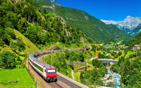 A red and white passenger train winds through a lush Alpine valley with mountain tunnels, a river, and snow-capped peaks in the background