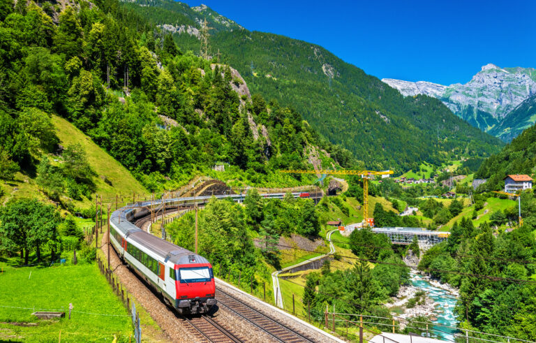 A red and white passenger train winds through a lush Alpine valley with mountain tunnels, a river, and snow-capped peaks in the background