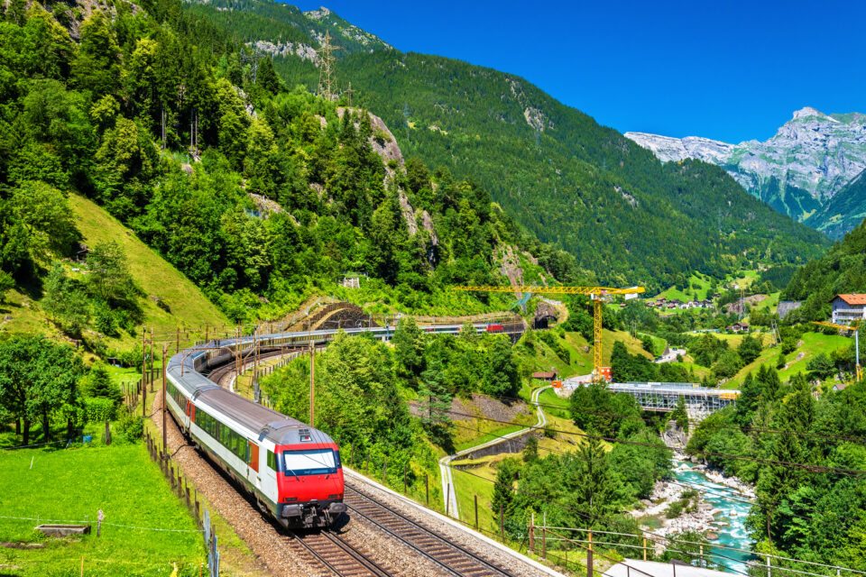 A red and white passenger train winds through a lush Alpine valley with mountain tunnels, a river, and snow-capped peaks in the background
