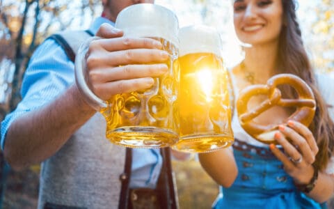 Two people in traditional Bavarian attire clinking large beer steins outdoors, with a woman holding a pretzel