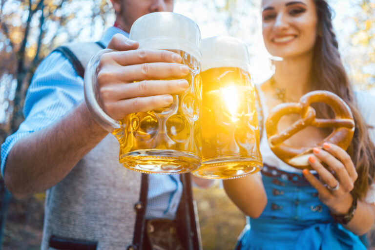 Two people in traditional Bavarian attire clinking large beer steins outdoors, with a woman holding a pretzel